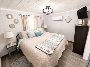 A bedroom with a bed, bedside tables, a lamp, a wall-mounted air conditioner, and a television on a stand.
