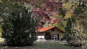 Eine kleine, einstöckige Holzhütte mit geneigtem Dach, umgeben von dichtem Grün und einer Rasenfläche.