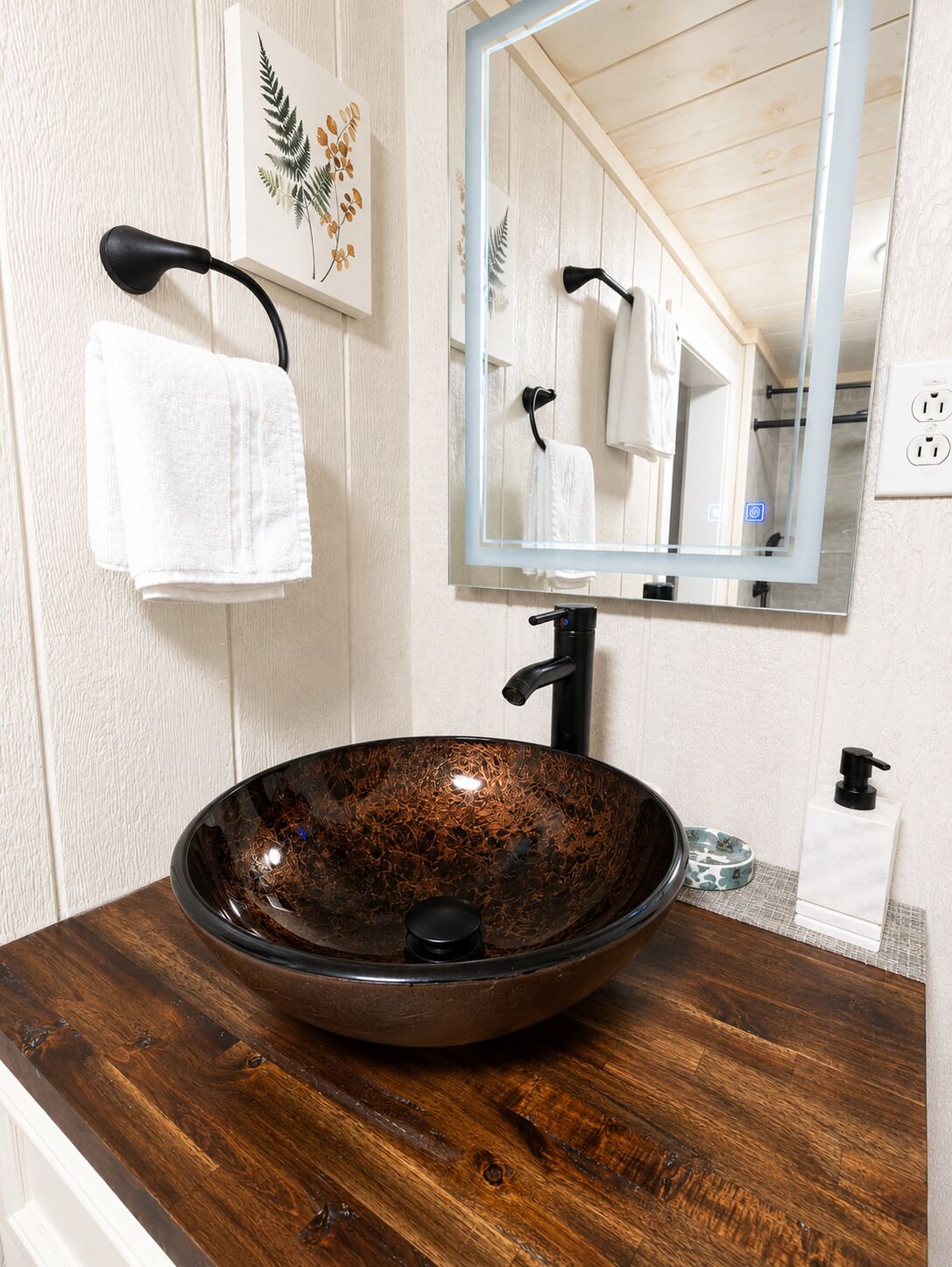 A bathroom with a wooden vanity, a vessel sink, a mirror, a towel rack, and a wall-mounted power outlet.