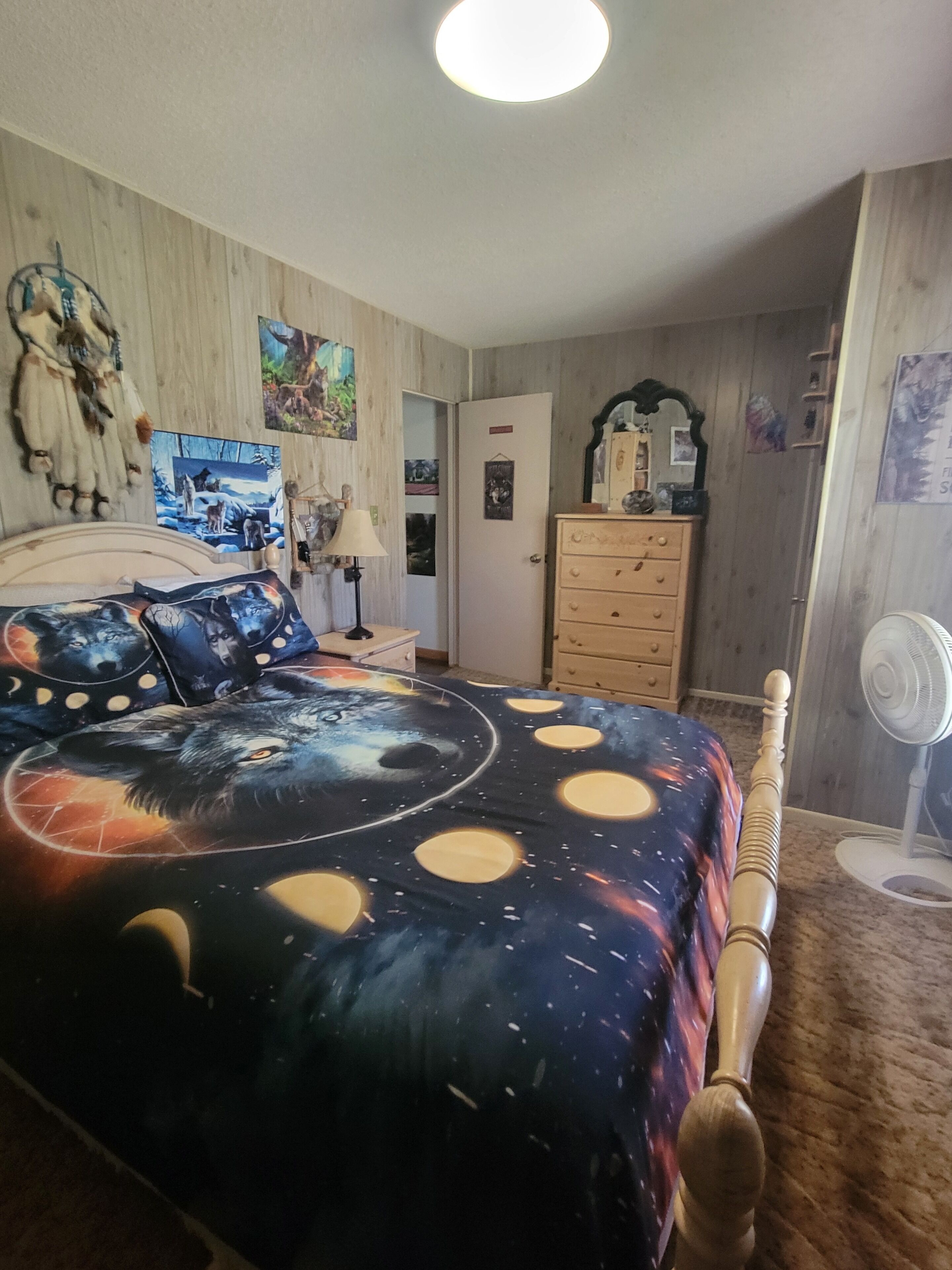 Une chambre à coucher avec un lit agrémenté d’une couvre-lit sur le thème de l’espace, une commode en bois, une table de chevet avec une lampe et un ventilateur fixé au mur.