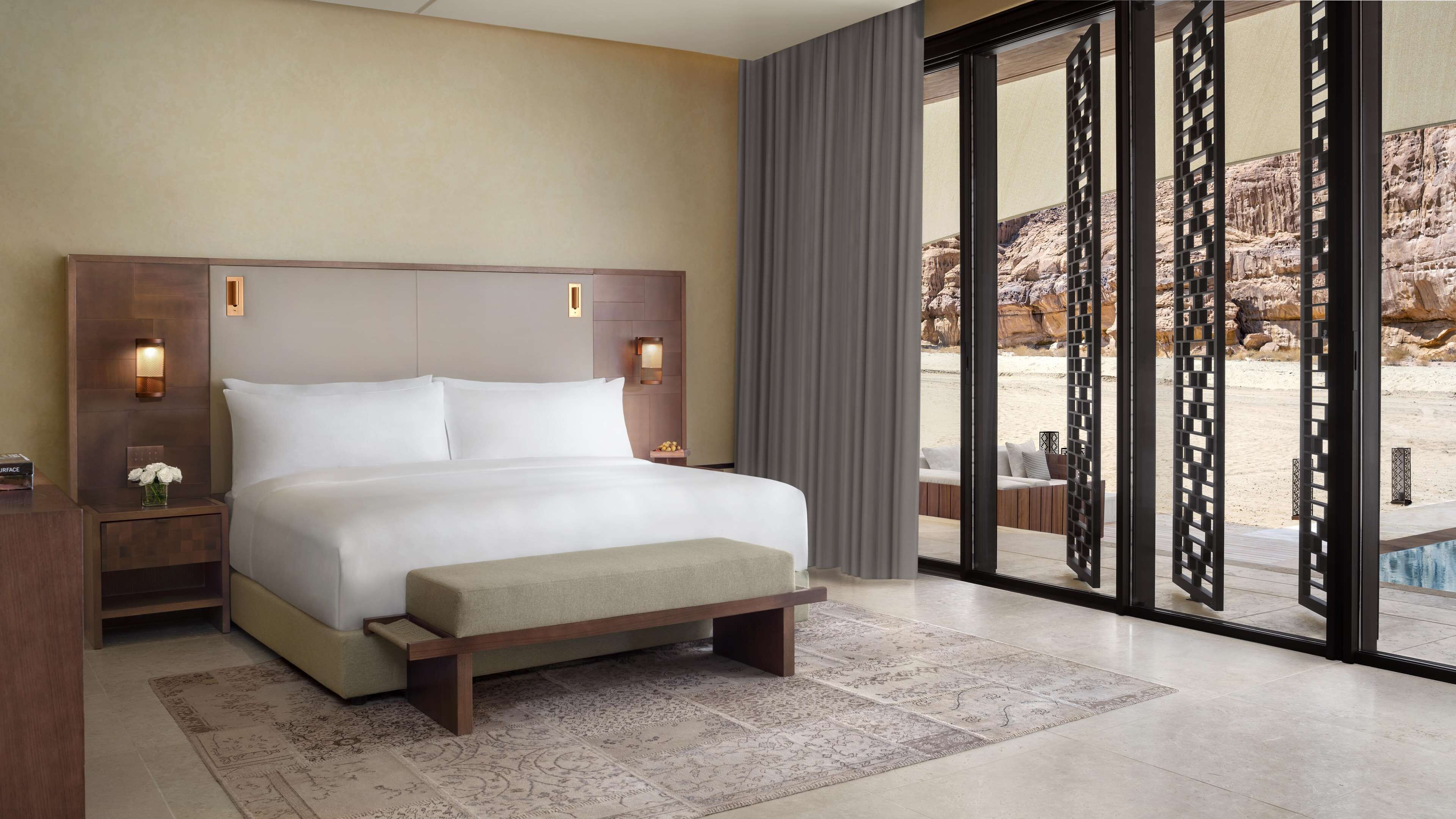 A hotel room with a large bed, a nightstand, a vase of flowers, and a view of a rocky landscape through a glass door.