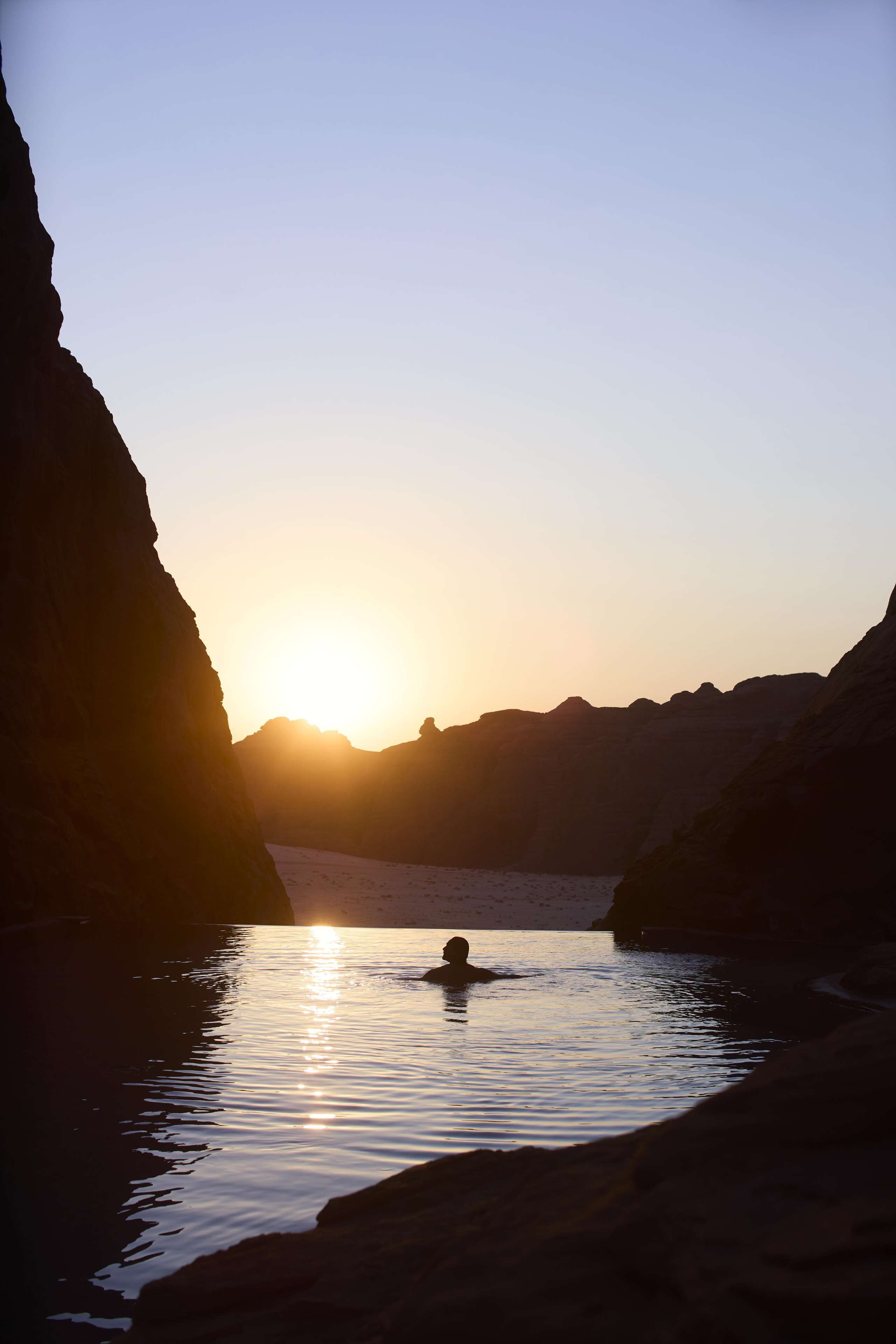 A person swimming in a natural water body surrounded by rock formations during sunset.
