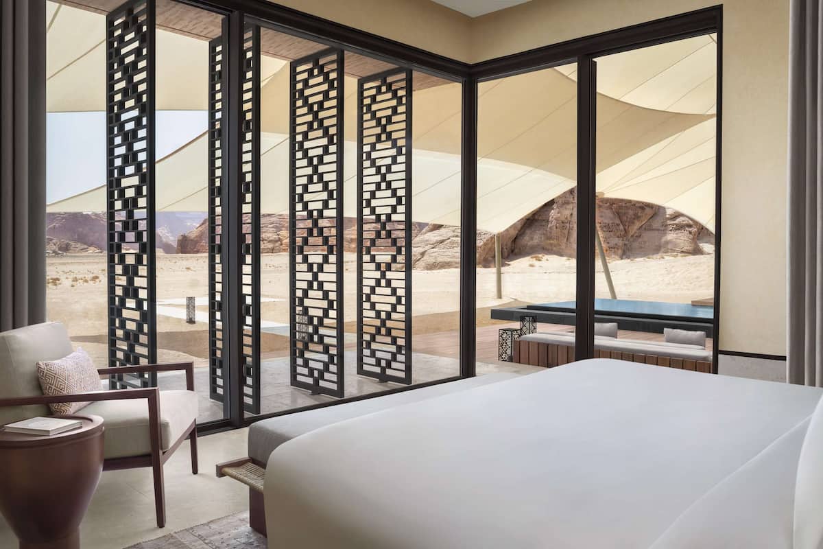 A hotel room with a large bed, a desk, a chair, and a view of a desert landscape through floor-to-ceiling windows.
