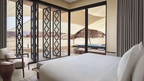 A hotel room with a large bed, a seating area with a table, and a view of a desert landscape through floor-to-ceiling windows. - Banyan Tree AlUla (Al-'Ula)