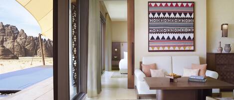 Habitación de hotel moderna con un amplio ventanal que ofrece vistas al desierto, una zona de estar acogedora con un sofá y una pequeña mesa, y un elemento decorativo en la pared.