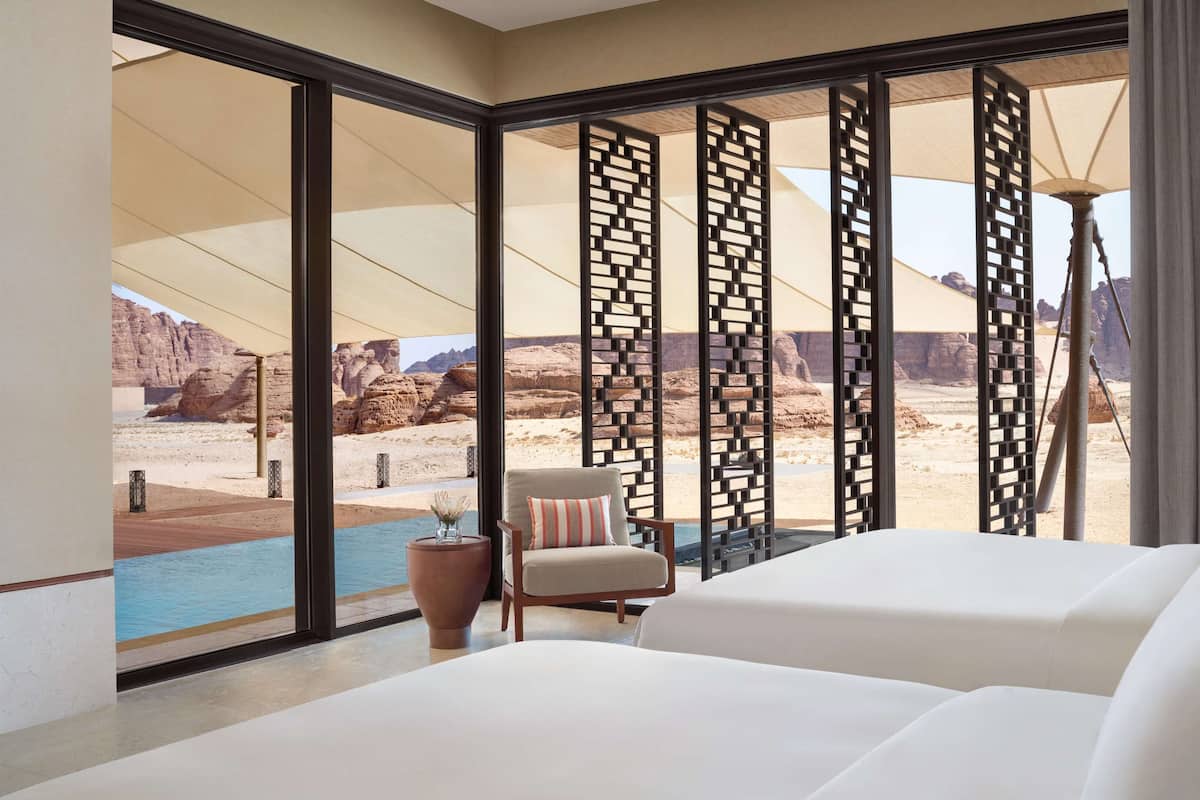 A hotel room with a large window, a bed, a chair, and a small table with a vase.