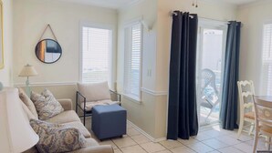 Ein Wohnzimmer mit Sofa, Hocker und Sesseln, einem Deckenventilator und Blick durch ein Fenster mit Vorhängen.