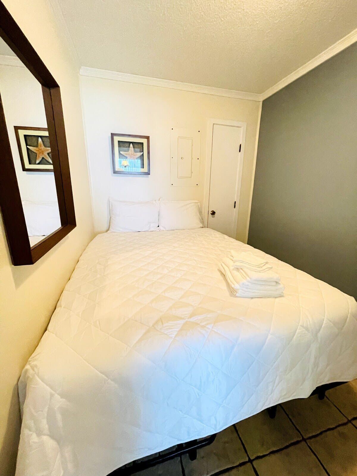 A neatly made bed with a white quilt in a room with a door and a framed picture on the wall.