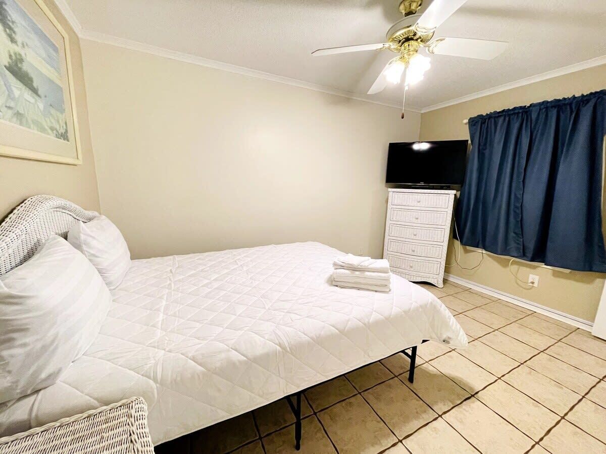 Een slaapkamer met een bed, een dressoir, een plafondventilator, een televisie en een raam met gordijnen.