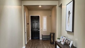 A hallway with a wooden floor, a black door, a bench, and a small table with framed pictures. - Purple Sage Garden Retreat (Las Cruces)