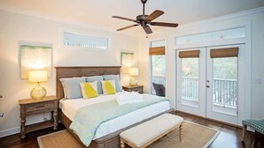 A bedroom with a wooden bed, two bedside tables with lamps, a ceiling fan, and a view of the outdoors through a door with a balcony. - Bird Song | Heated Pool | 4 Bikes | Dog Friendly (Santa Rosa Beach)
