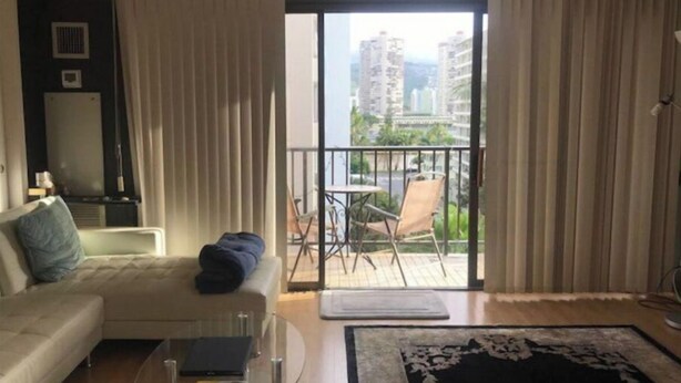 A modern living room with a sofa, a glass coffee table, and a view of a balcony with outdoor furniture. - Aloha Waikiki! (Honolulu)