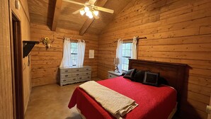 Een slaapkamer in een houten hutje met een bed, een dressoir en een plafondventilator.