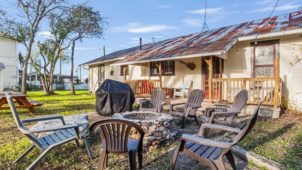 A rustic outdoor seating area with a fire pit, surrounded by Adirondack chairs, in front of a house with a weathered metal roof. - Carriage Inn - Cottage (Bandera)
