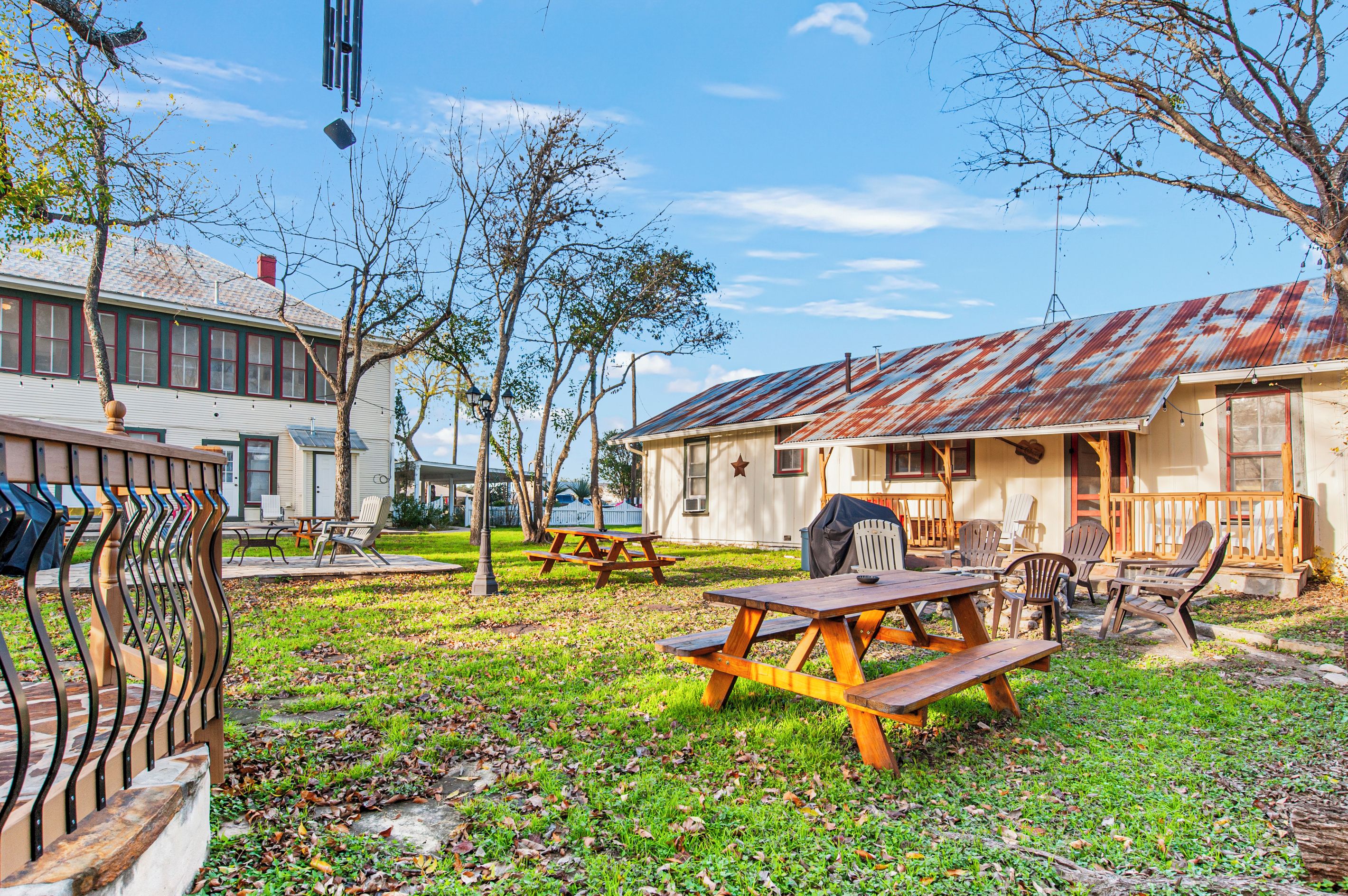 A picnic area with wooden benches and tables, surrounded by trees and a building with a rusted roof.