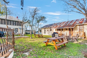 A picnic area with wooden benches and tables, surrounded by trees and a building with a rusted roof. - Carriage Inn - Cottage (Bandera)