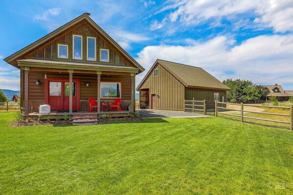A wooden cabin with a porch and red door, surrounded by a fenced yard.