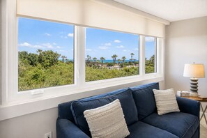 A room with a blue sofa, two white pillows, a lamp, and a view of a beach and palm trees through large windows. - Oceanview 4BR condo in Amelia Island Plantation with pool, pet friendly! (Fernandina Beach)