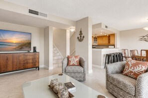 A modern living room with a flat-screen TV, a wooden entertainment unit, a staircase, and a kitchen area with wooden cabinets. - Oceanview 4BR condo in Amelia Island Plantation with pool, pet friendly! (Fernandina Beach)