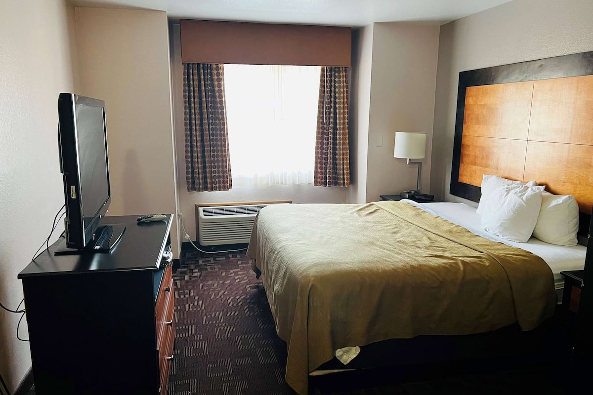 A hotel room with a bed, a desk with a television, an air conditioner, and a window with curtains.