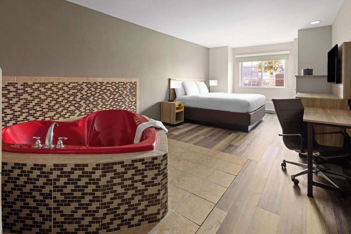 A hotel room with a large red jacuzzi, a bed, a desk with a chair, and a TV mounted on the wall.