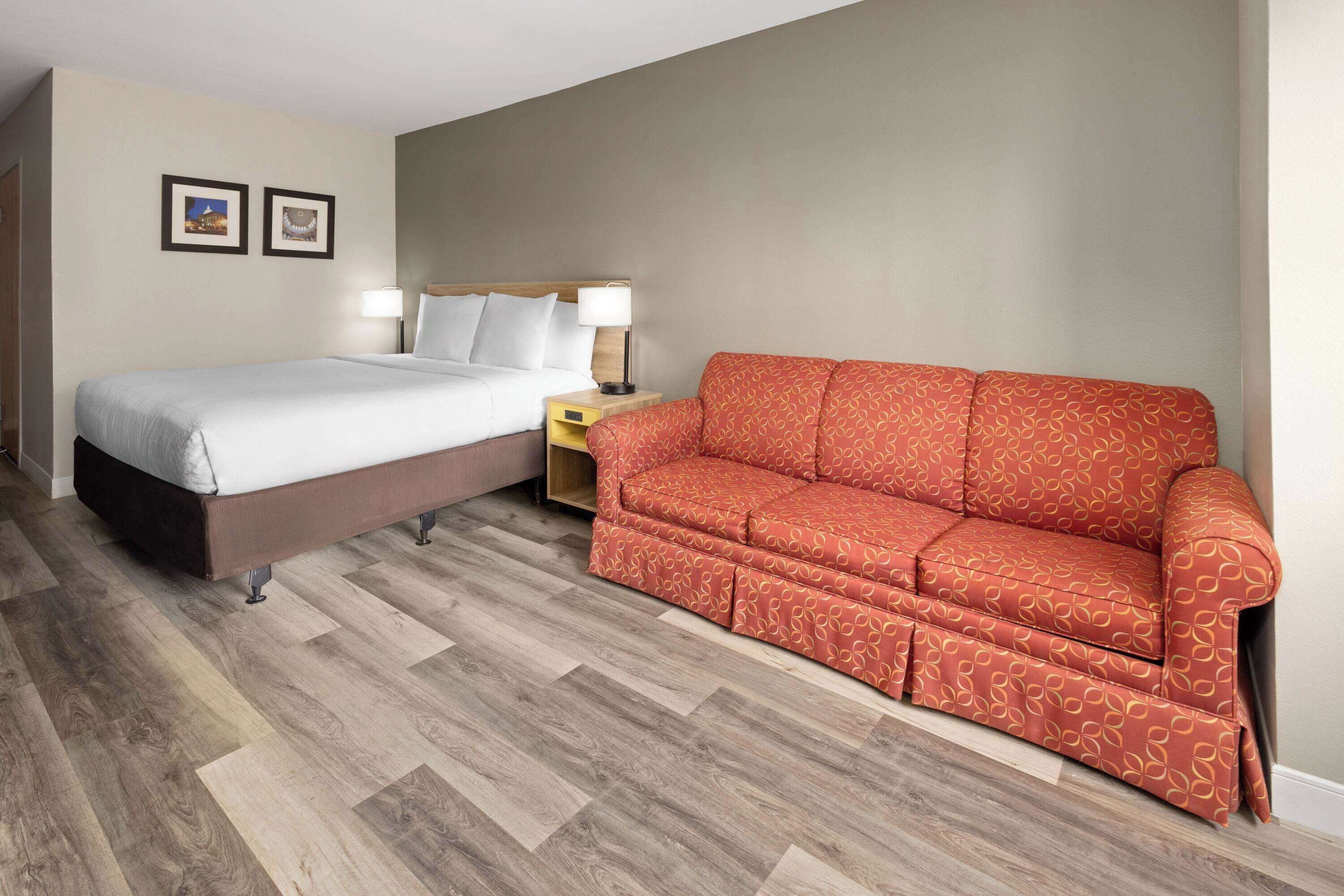 Quarto de hotel com cama, sofá vermelho estampado, mesa de cabeceira com abajur e duas imagens emolduradas na parede.