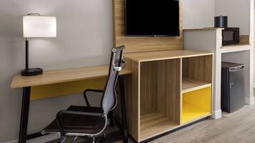 A modern office space with a wooden desk, a black office chair, a mounted flat-screen TV, a lamp, and a small black refrigerator.
