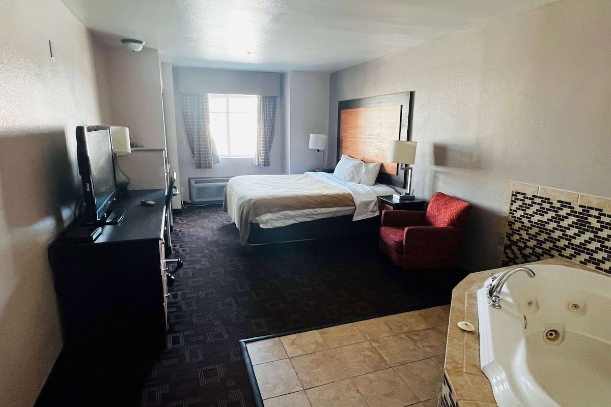 A hotel room with a bed, a desk with a computer, a chair, a bathroom with a bathtub, and a window with curtains.