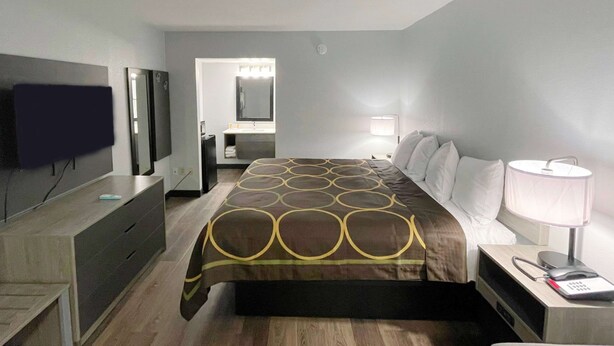 Habitación de hotel con cama, mesita de noche con lámpara, teléfono, televisor montado en la pared y baño a la vista a través de una puerta abierta.