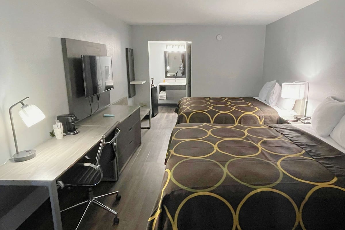 A hotel room with a bed, a desk with a chair, a TV, a mirror, and a bathroom with a sink and mirror.