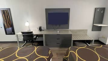 Habitación de hotel con escritorio, silla, cama con ropa de cama estampada, televisor montado en la pared y una pequeña estantería.