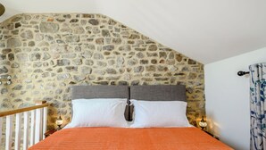 Un dormitorio con pared de piedra, cabecera de madera, cama con ropa de cama blanca y dos lámparas de noche.