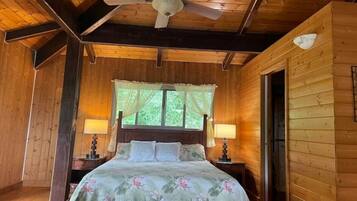 Dormitorio de cabaña de madera con cama, dos mesitas de noche, ventilador de techo y ventana con cortinas.