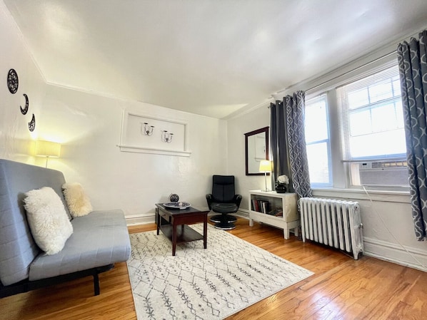A living room with a grey sofa, a small coffee table, a black armchair, a white side table, a mirror, and a radiator.