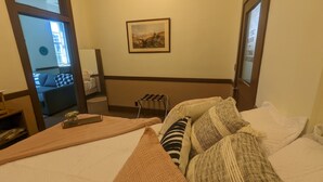Hotellihuone, jossa on sänky, puinen yöpöytä, pieni pöytä maljakolla, seinällä oleva kehystetty kuva sekä näkymä käytävään ja toiseen huoneeseen.