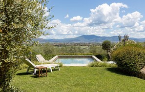 Zona de piscina con dos sillones de descanso y una mesita, rodeada de vegetación y vistas a un paisaje urbano y montañas.
