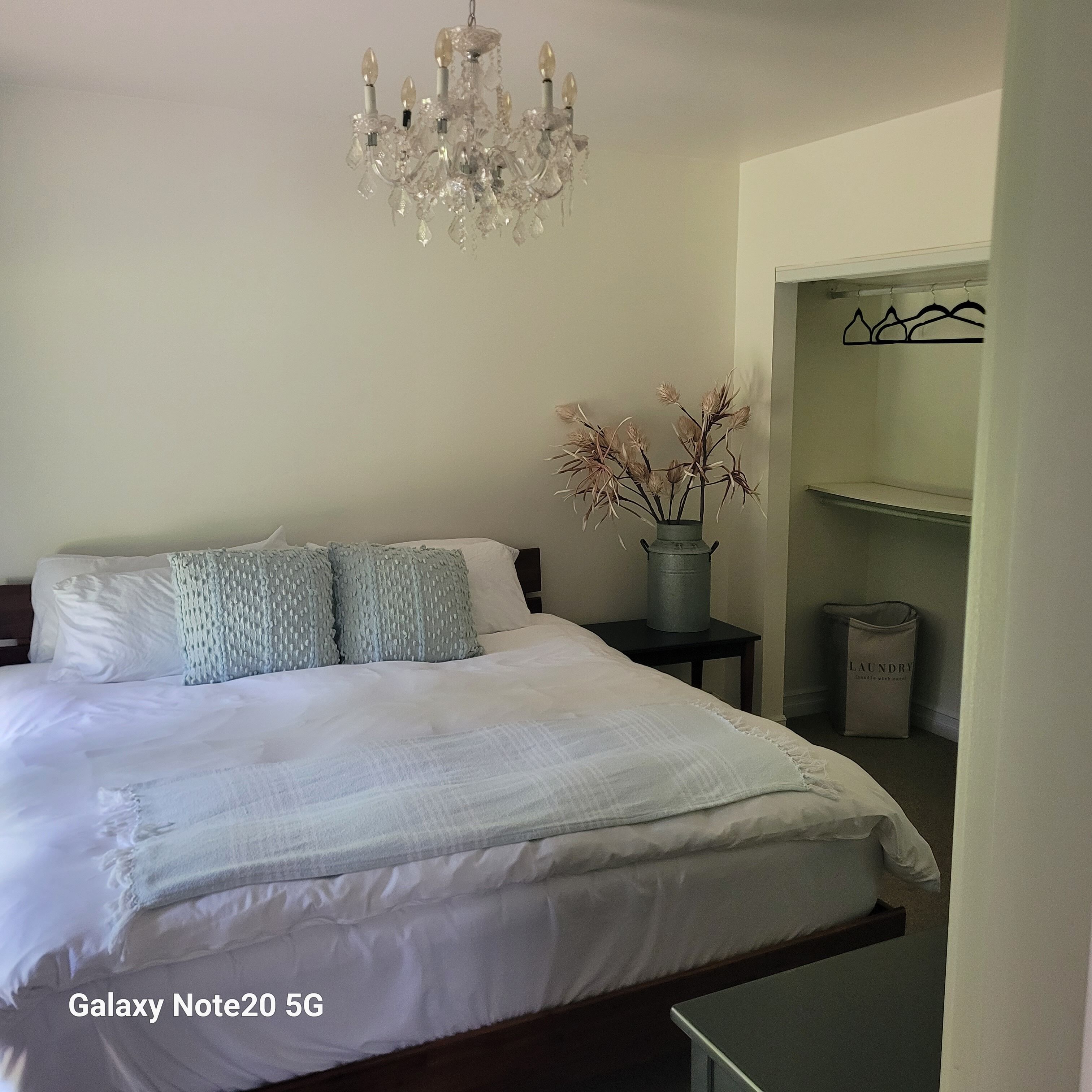 A neatly made bed with a Galaxy Note20 5G, a chandelier, and a vase with dried flowers.