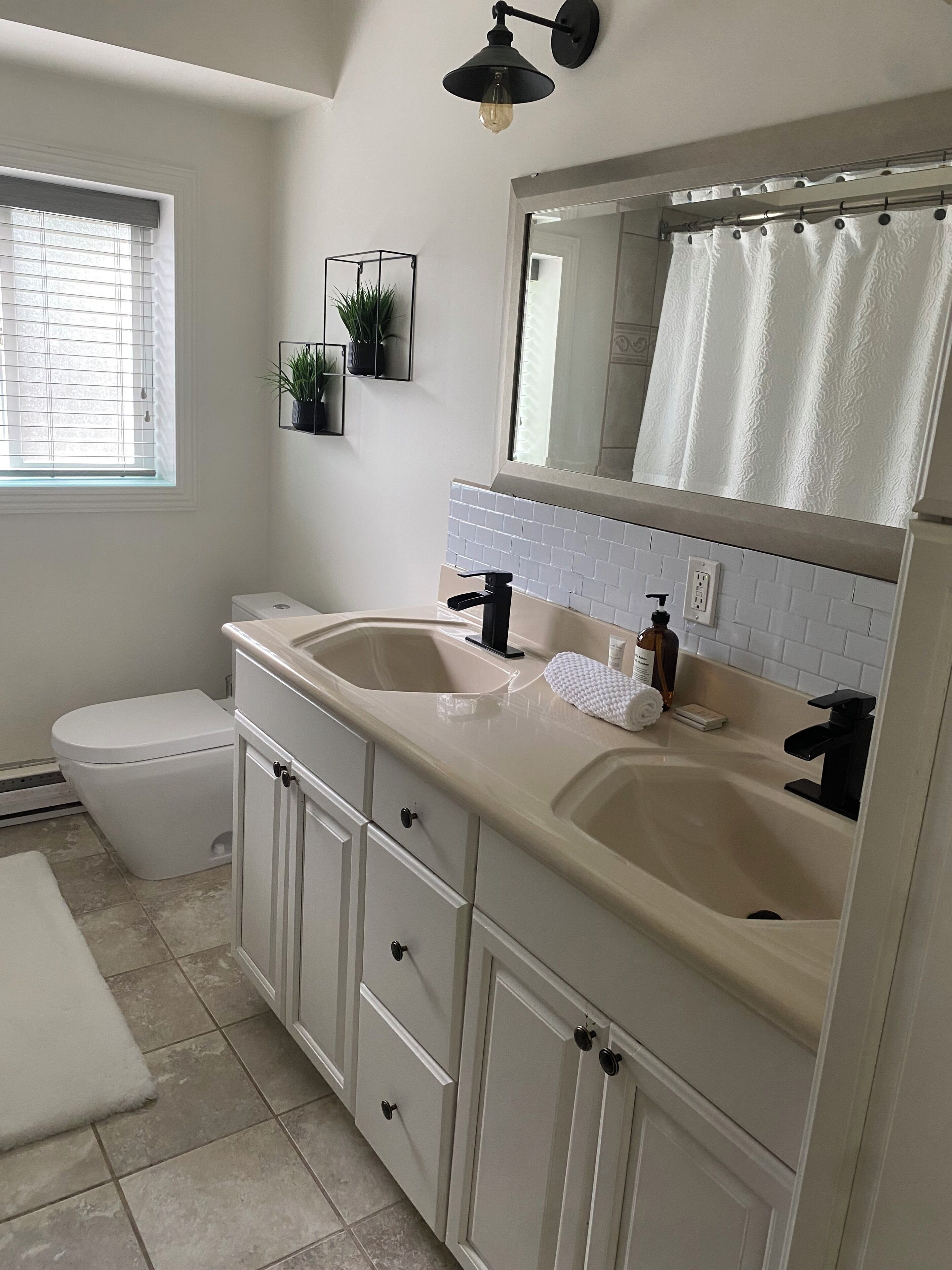 A bathroom with two sinks, a toilet, a window with blinds, a towel rack, and a mirror with a towel hanging.