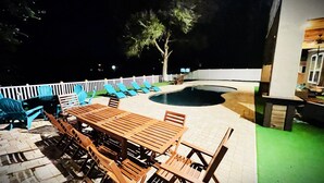 An outdoor patio with a pool, wooden furniture, and a white fence.