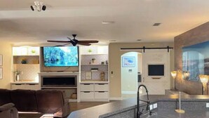 A modern living room with a large TV, a ceiling fan, and a kitchen area with a sink and countertop.