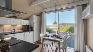 Una cocina moderna con gabinetes blancos, encimera negra y una zona de comedor con mesa y sillas. Un ventanal ofrece vistas a un paisaje natural.