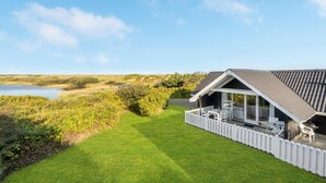 Una casa moderna con un amplio ventanal, una cerca de piquetes blancos y vistas a un lago y dunas.