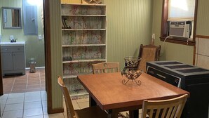 Un coin repas avec une table et des chaises en bois, une bibliothèque et un climatiseur d’époque.