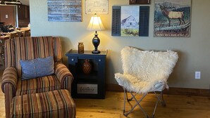Un salon avec un fauteuil à rayures, un fauteuil blanc et moelleux, une petite table d’appoint avec une lampe, et divers éléments de décoration murale, dont une enseigne sur laquelle on peut lire : « TOUS LES CHASSEURS, LES PÊCHEURS ET TOUS LES AUTRES MENTEURS SONT LES BIENVENUS ».