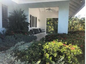 Un área de estar exterior moderna con una estructura blanca, rodeada de abundante follaje y plantas en flor.