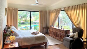 A spacious bedroom with a large bed, a sitting area with a chair, a wooden cabinet, and a view of a swimming pool.