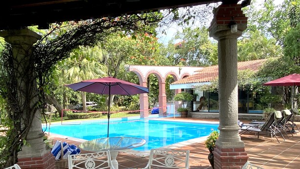 A pool area with a glass table and chairs, surrounded by brick pillars and covered by umbrellas.
