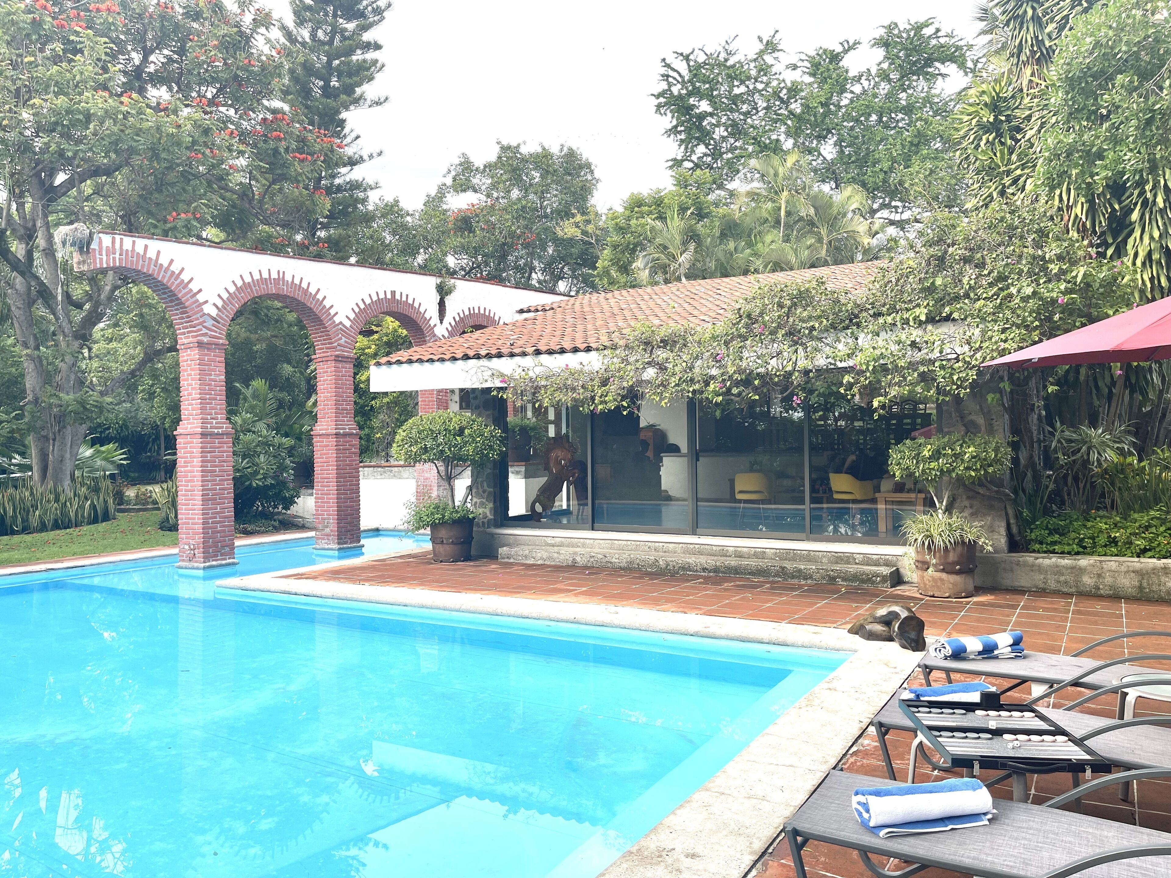 A swimming pool with a brick archway, a covered patio area with seating, and lush greenery in the background.