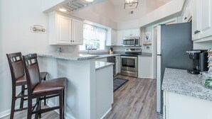 Una cocina moderna con armarios blancos, encimeras de granito, una isla central con taburetes y electrodomésticos de acero inoxidable, y vistas al exterior.