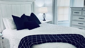 Un dormitorio con cama blanca, almohadas oscuras, un plaid estampado, una lámpara de noche, un aparador y una ventana con persianas.